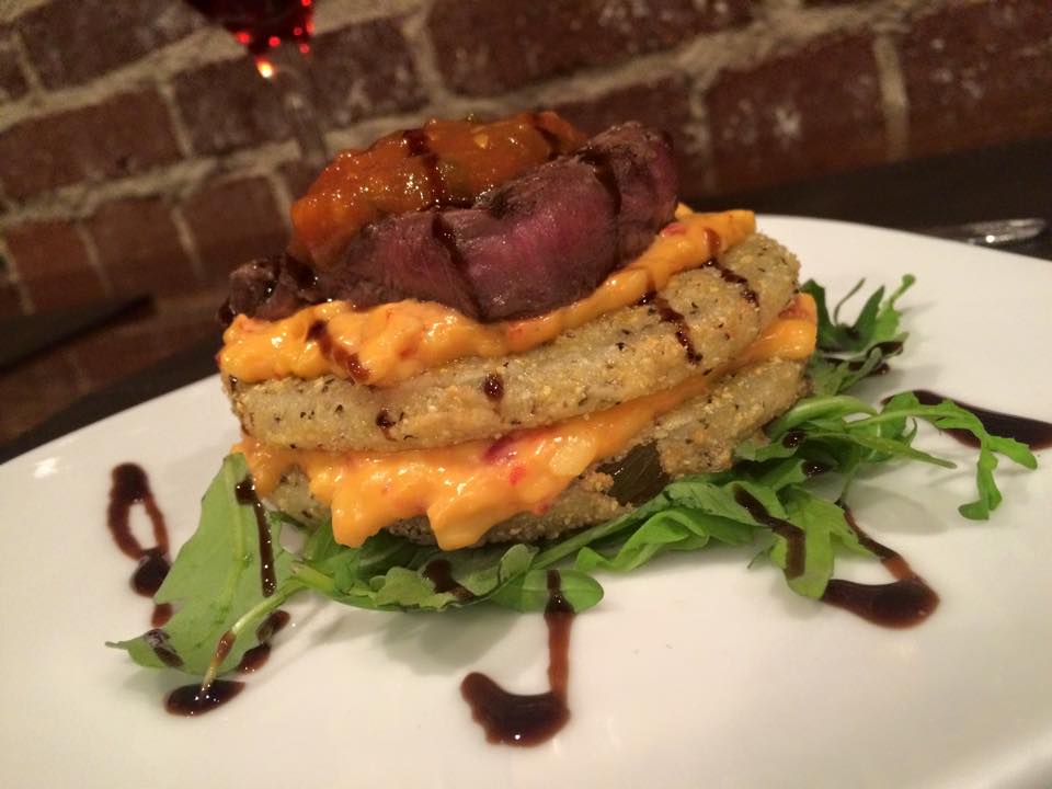 Petite Filet Mignon sits atop fried green tomatoes covered in local, stone-ground cornmeal, pimento cheese with a tomato jam and a balsamic glaze, sits on a bright white plate against a brick wall background.